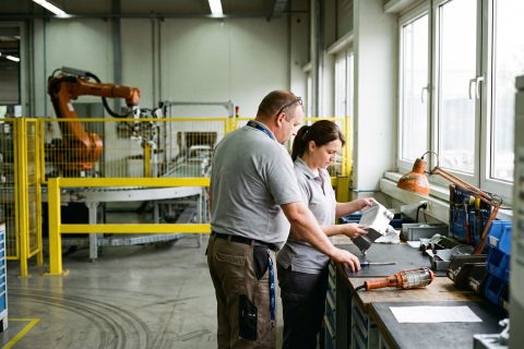 Zwei Beschäftigte begutachten ein Aluminiumgehäuse an einer Werkbank, dahinter eine Roboterzelle.