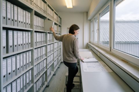 Mitarbeiterin zieht einen Ordner aus einer Wand mit gleichförmigen Aktenordnern.