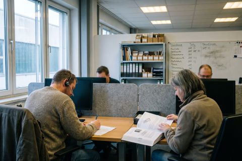 Kundenservice-Team an benachbarten Schreibtischen, im Hintergrund ein Regal mit Handbüchern.