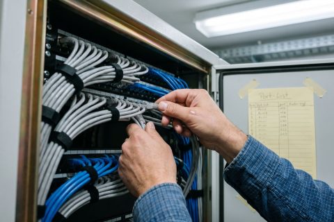Hände legen Patchkabel in einem geöffneten Netzwerkschrank, daneben eine handschriftliche Portliste.