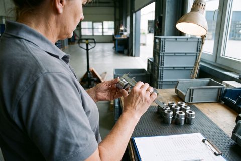 Mitarbeiterin prüft ein Drehteil mit dem Messschieber am Wareneingang, daneben eine Prüfliste.